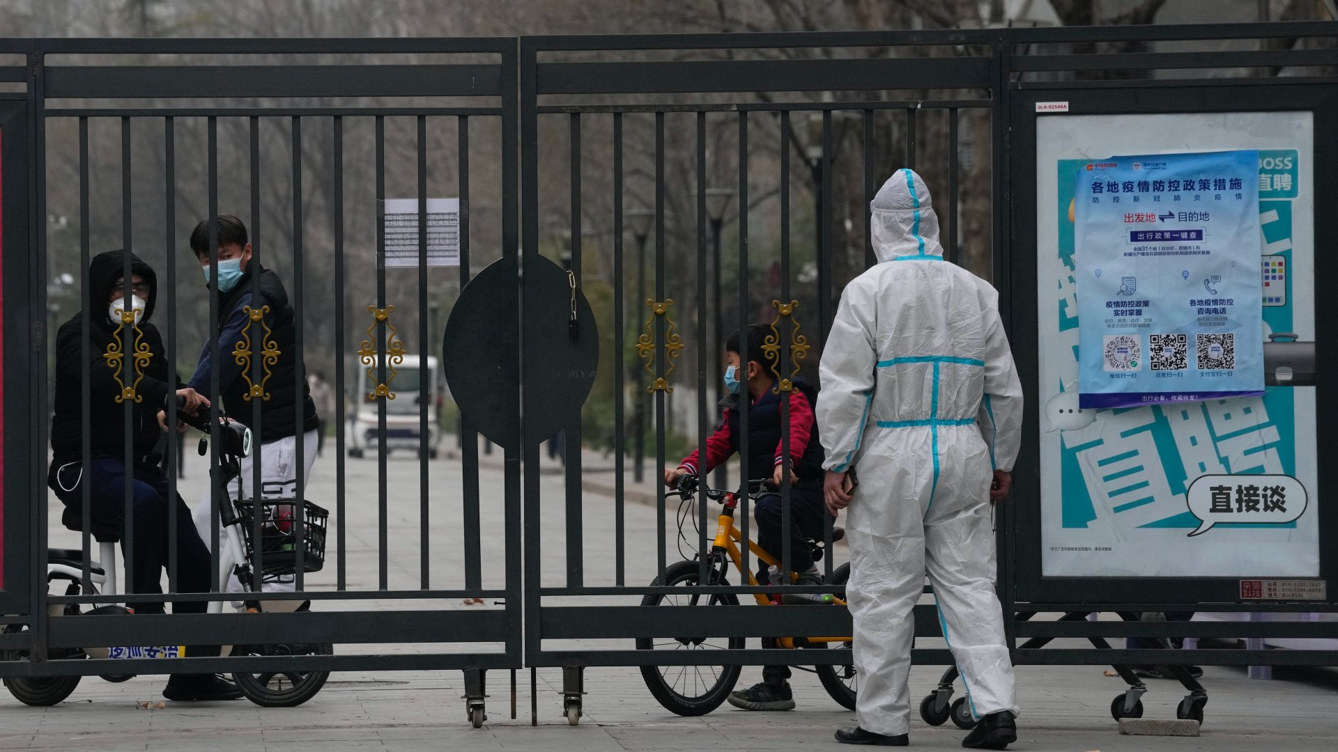 Egy lezárt lakóközösség tagjai egy védõöltözetet viselõ munkással az elõtérben Pekingben 2022. március 13-án