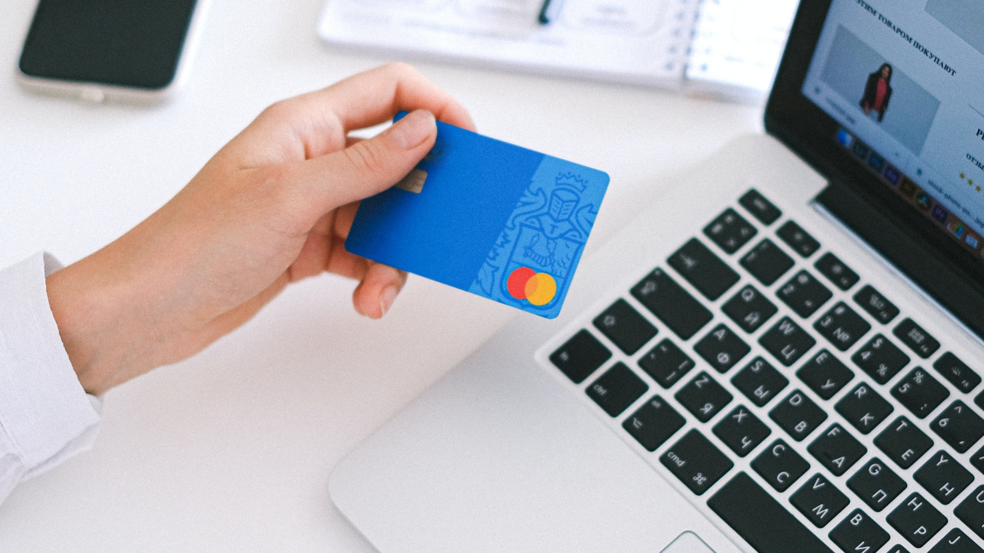 Internetes vásárláshoz használt bankkártyát fog a kezében a laptopnál ülő nő