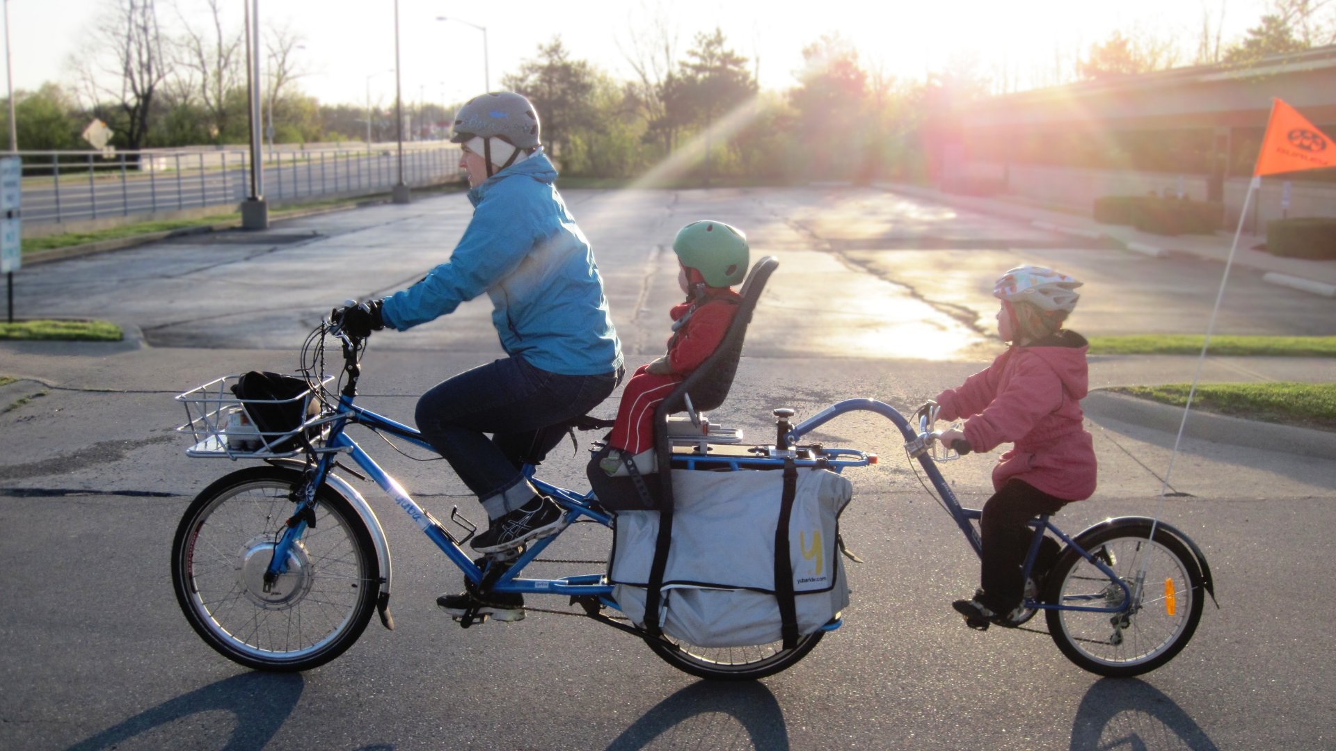Bicikliző szülő két gyerekkel a gyerekülésben és egy hozzákötött biciklin