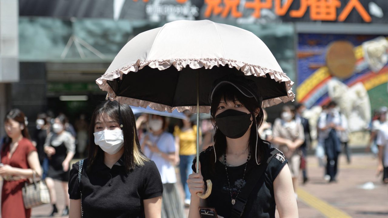 japán lány napernyő alatt, maszkban sétál a nyüzsgő utcán.
