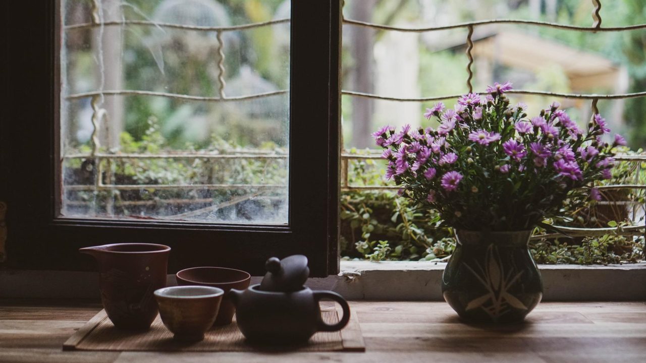 Egy ablak előtt lila virágoka tartalmazó váza és néhány teás csésze látható.