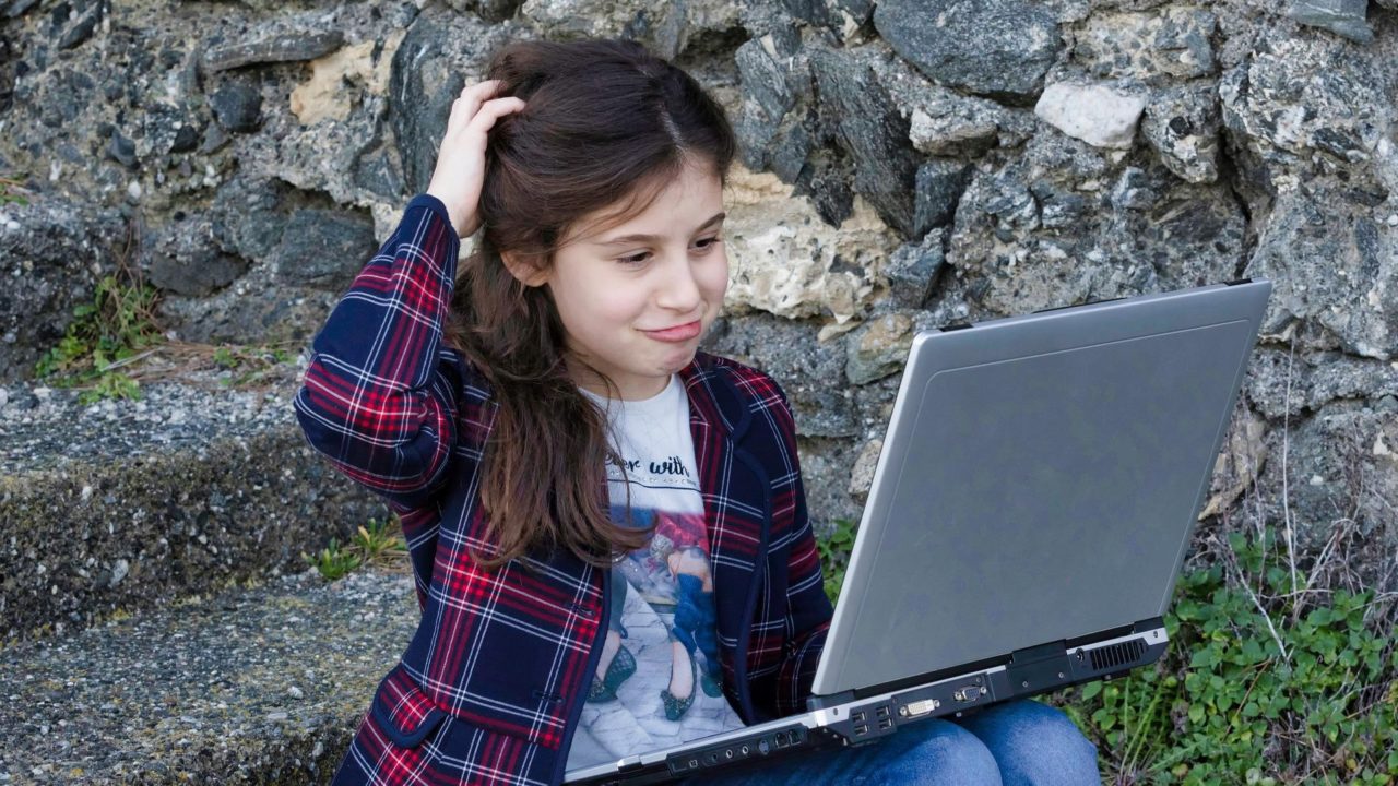 Kislány vakarja a fejét egy laptoppal az ölében.