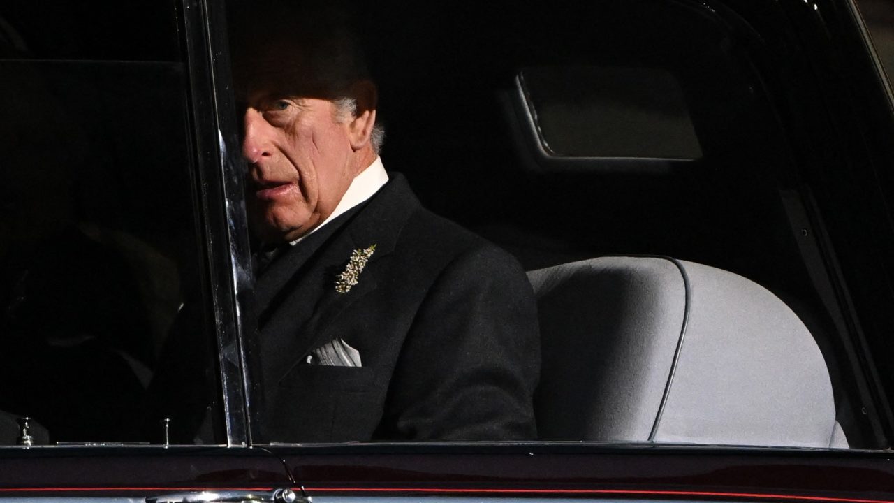 III. Károly király egy autóból néz kifelé. Az arca félig árnyékban van.