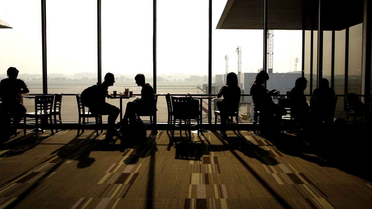 Repülőtéri terminálon asztaloknál cseveg emberek.