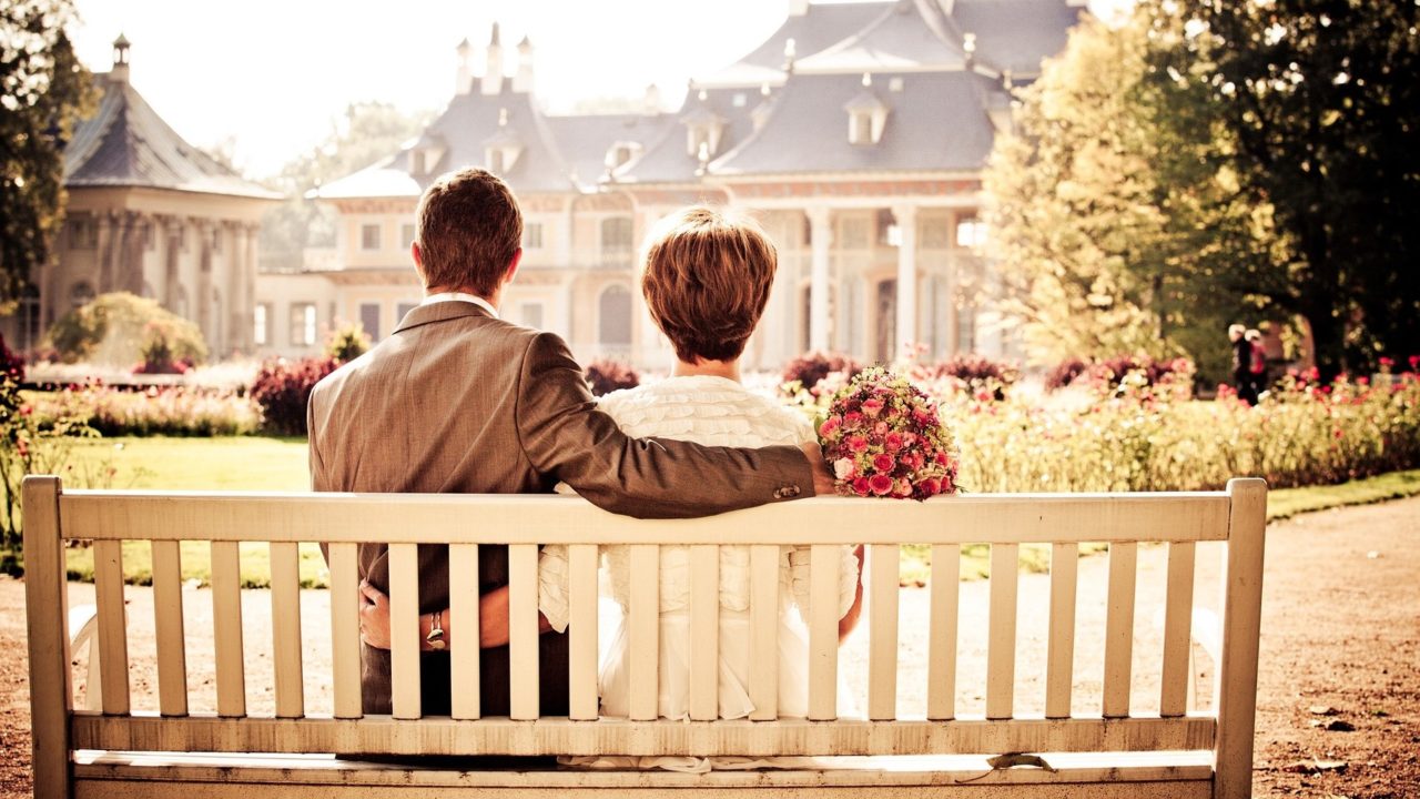 Egy férfi és egy nő ül egy padon háttal nekünk. Feltehetően menyasszony és férj.