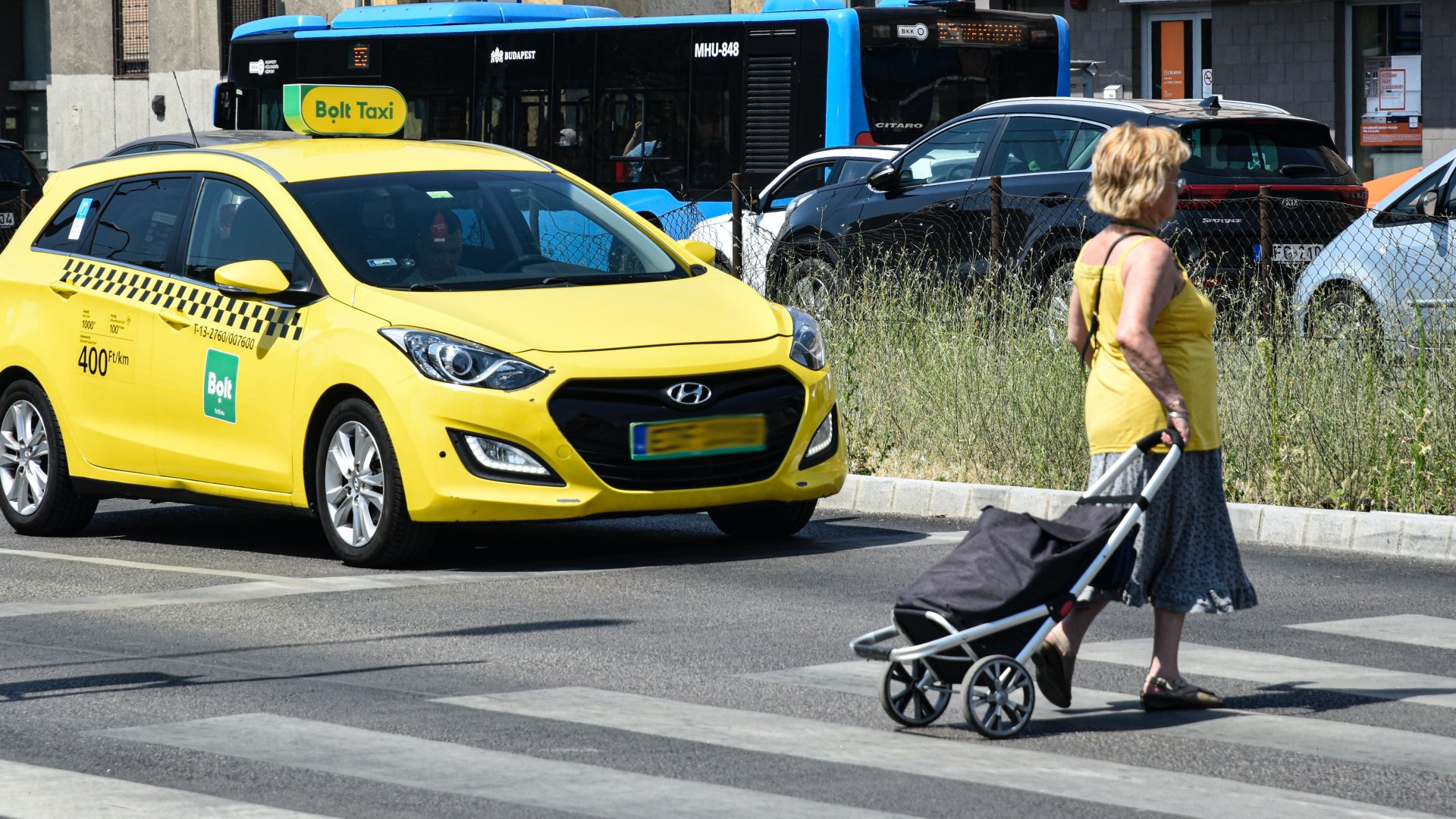 Nõ gurulós bevásárlótáskát maga után húzva megy át a gyalogátkelõhelyen, egy az áthaladását biztosító Bolt piacvezetõ európai utasszállító alkalmazás taxi gépkocsija elõtt a zuglói Bosnyák téren