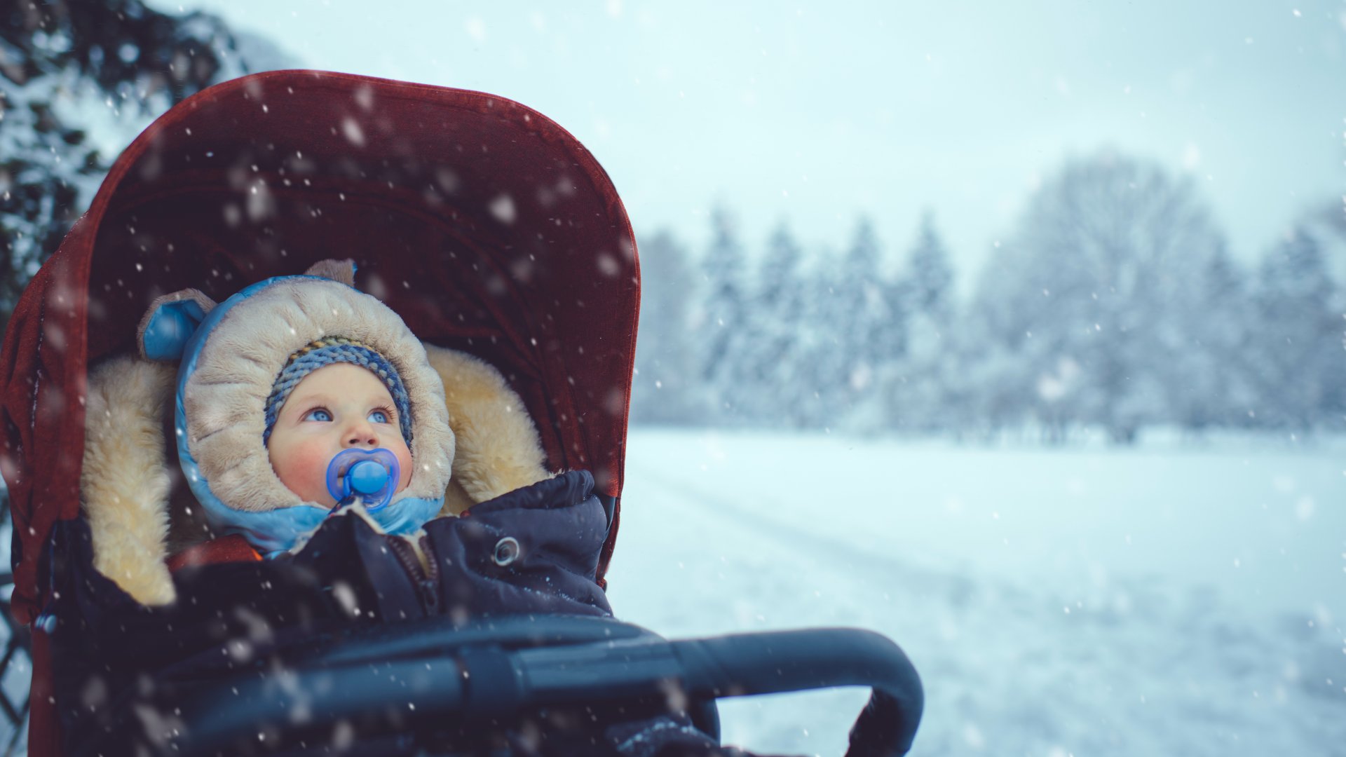 Egy babakocsiban ülő gyerek vastag ruhában figyeli a hóesést
