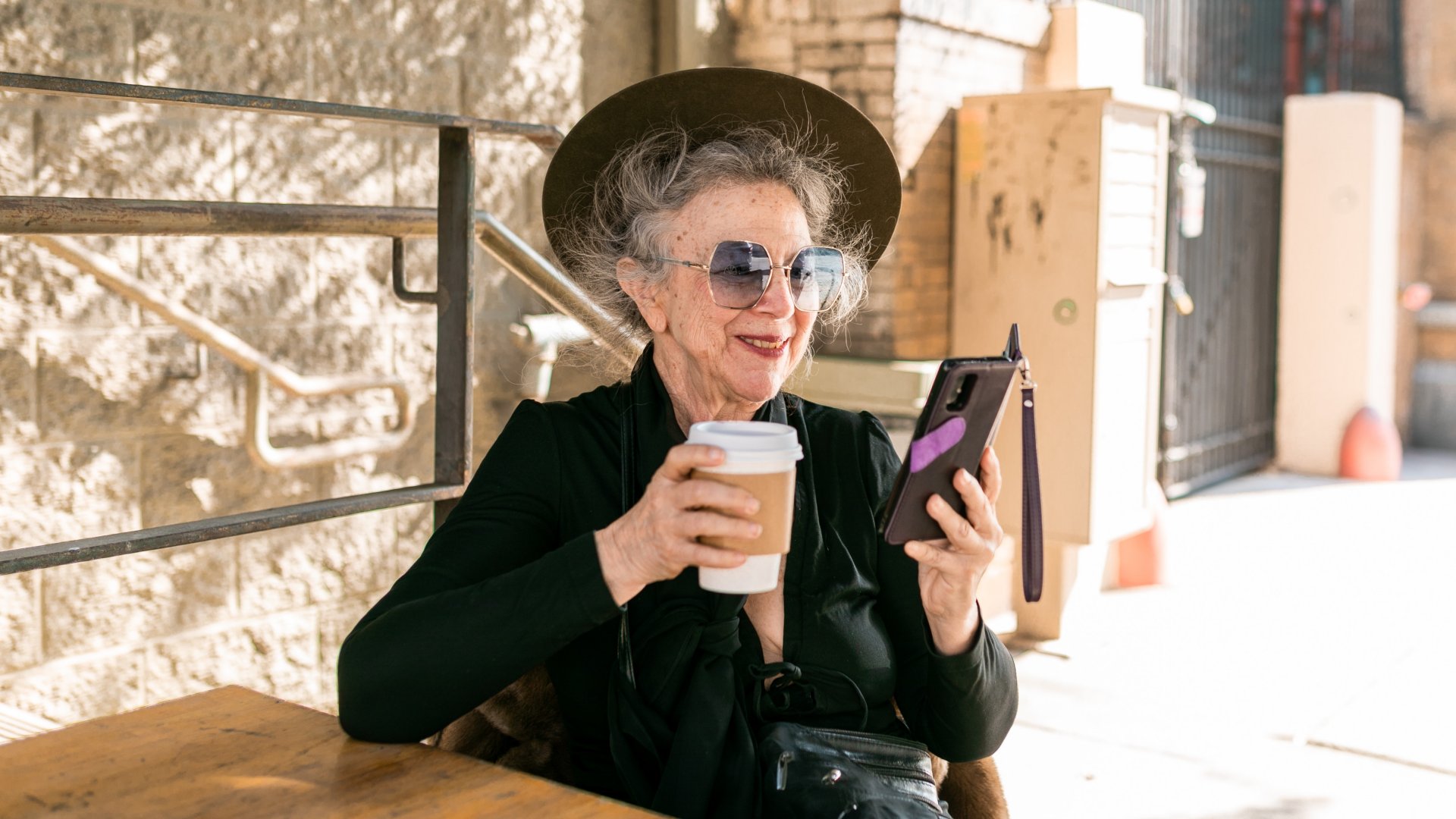 Nyugdíjas nő ül egy kávézó kinti asztalánál és a telefonját nézi