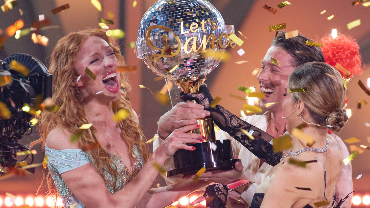 Anna Ermakova modell és profi táncpartnere, Valentin Lusin a műsorvezető Victoria Swarovski mellett a trófeával a kezükben ujjonganak a győzelemről az RTL Let's Dance című élő táncműsorának döntője után