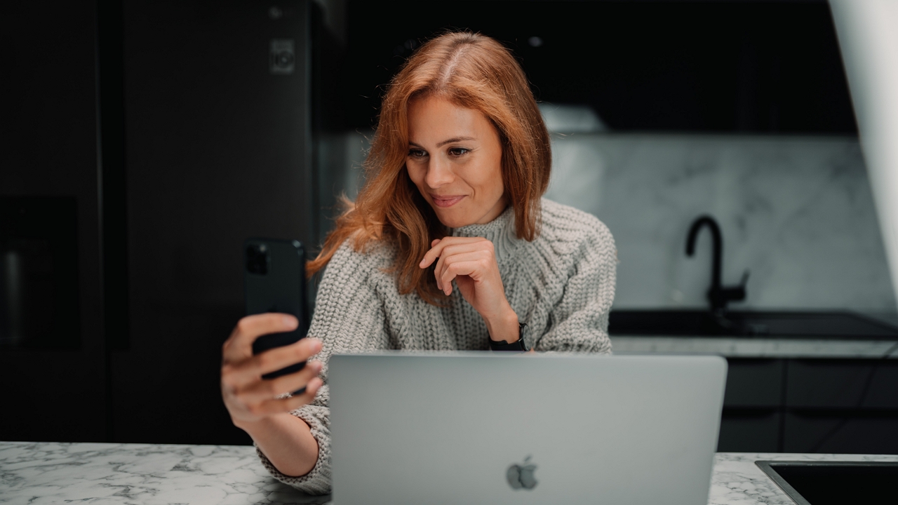 egy nő fotózza magát telefonnal, egy laptop előtt
