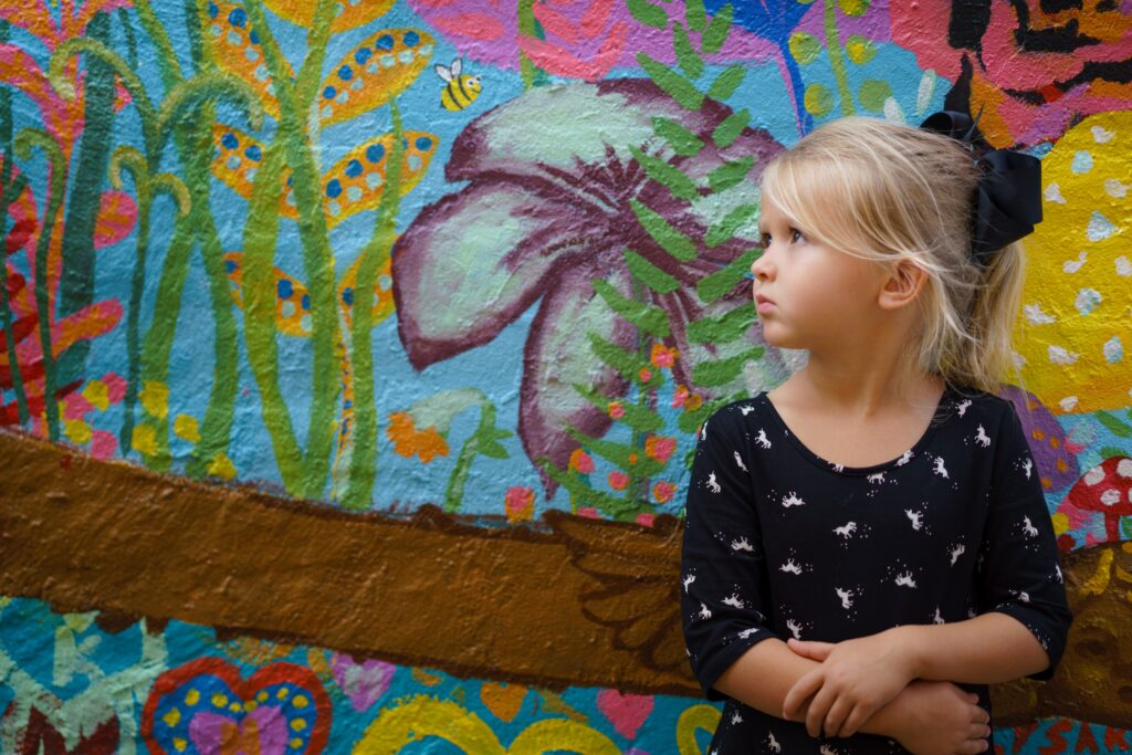 Szőke kislány áll egy színes fal előtt, hajában fekete masni.