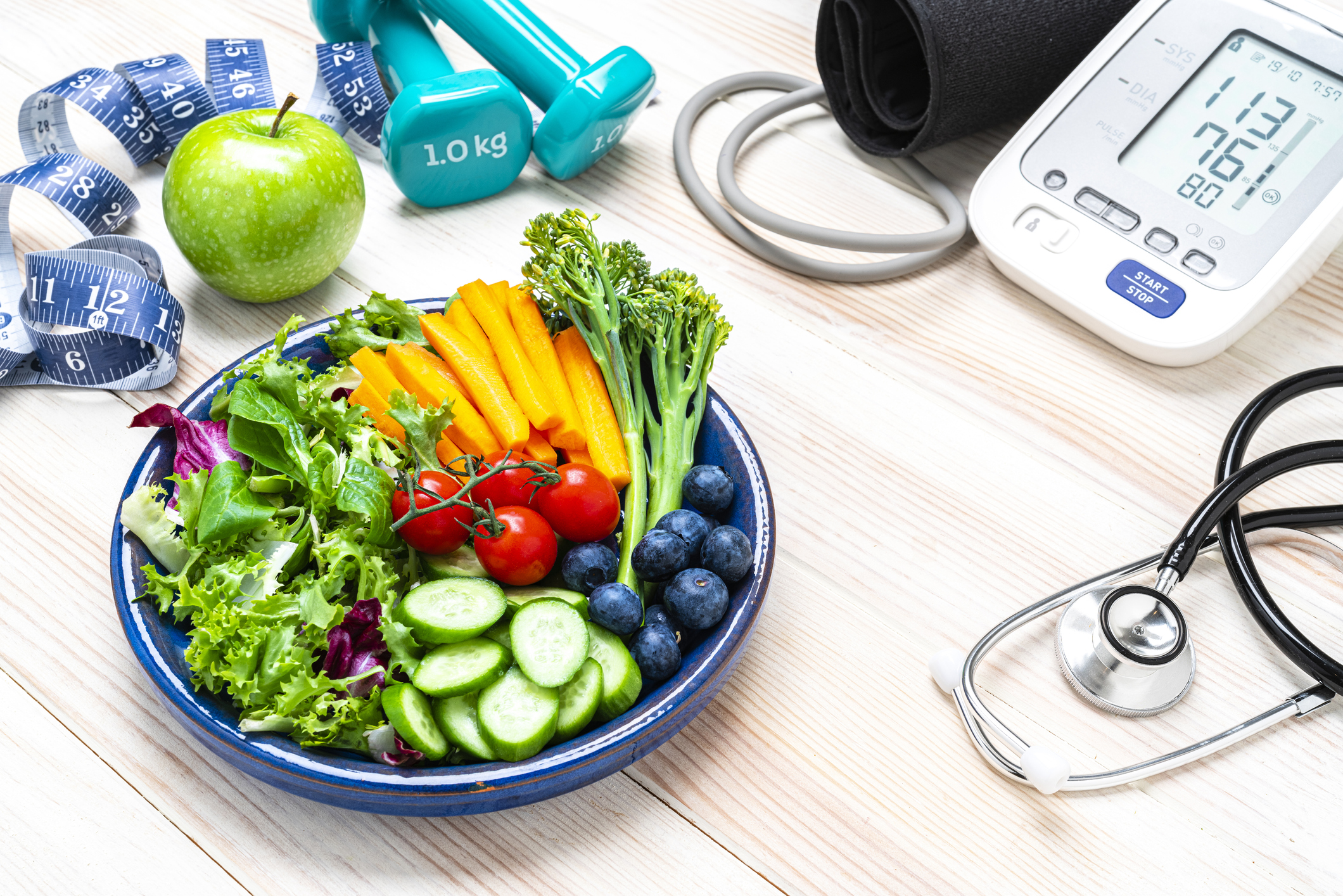 Egy tál zöldség hever az asztalon. Mellette egy vérnyomásmérő és edzéshez való súlyok vannak odakészítve.