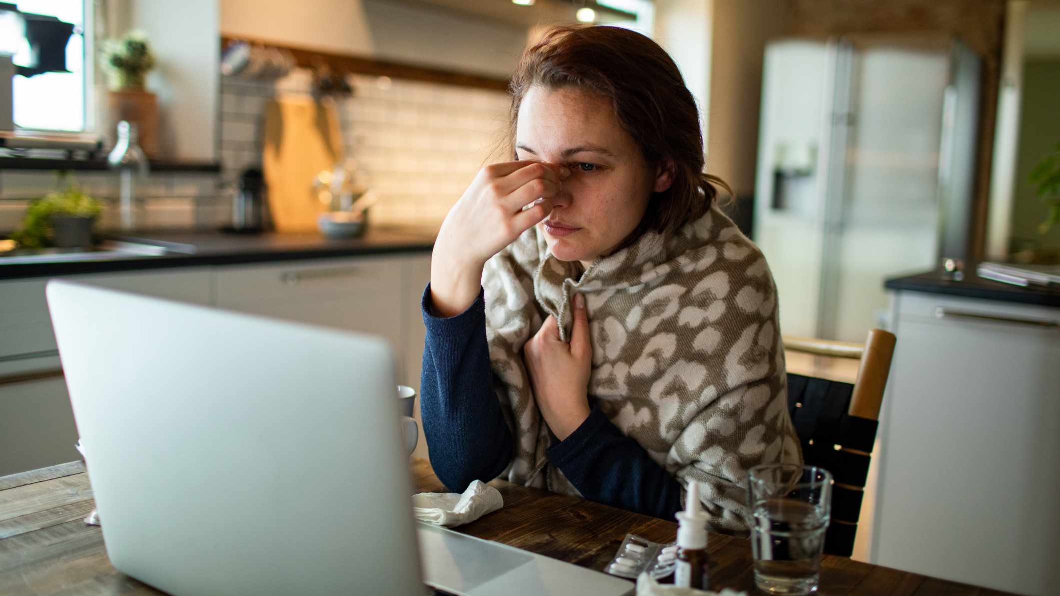 Egy fiatal nő ül az asztalánál, miközben dolgozik a laptopján. Fájdalja az orra körüli részt, az asztalon orrspray és zsepi hever.