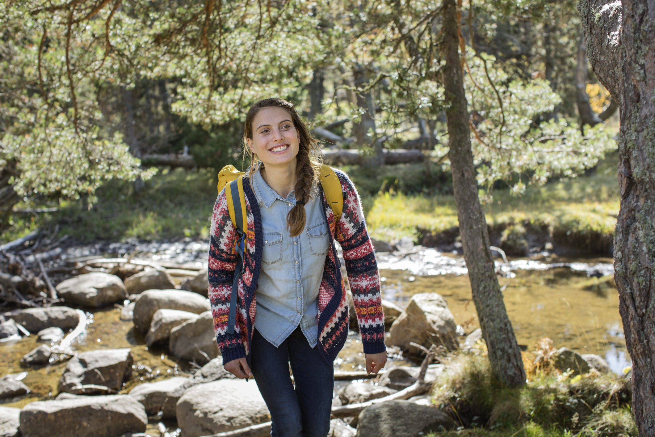 Egy fiatal nő túrázik az erdőben, közben mosolyog. A háttérben fák és egy patak lászik.