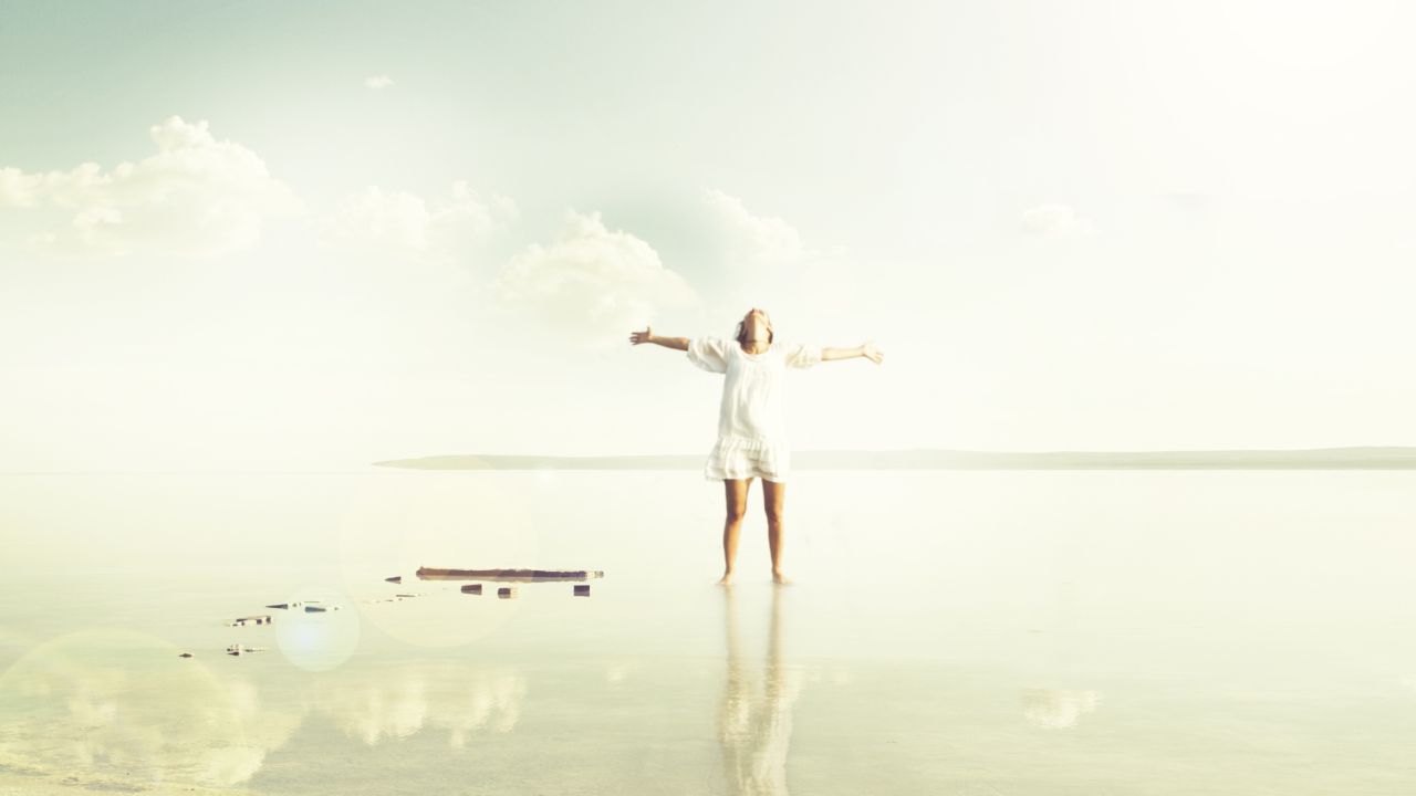 Fehér ruhás nő tárt karokkal áll a vízparton
