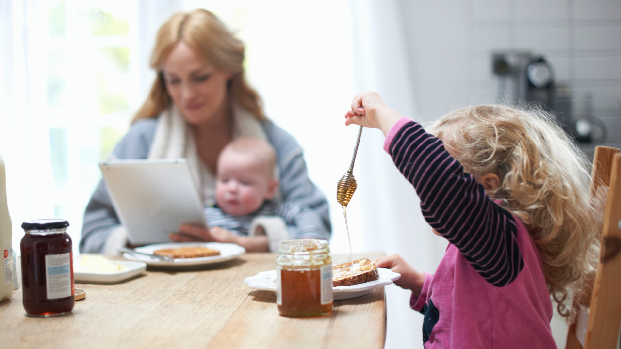 Kislány próbál mézet enni a reggelizőasztalnál, háttérben anyuka kisbabával.