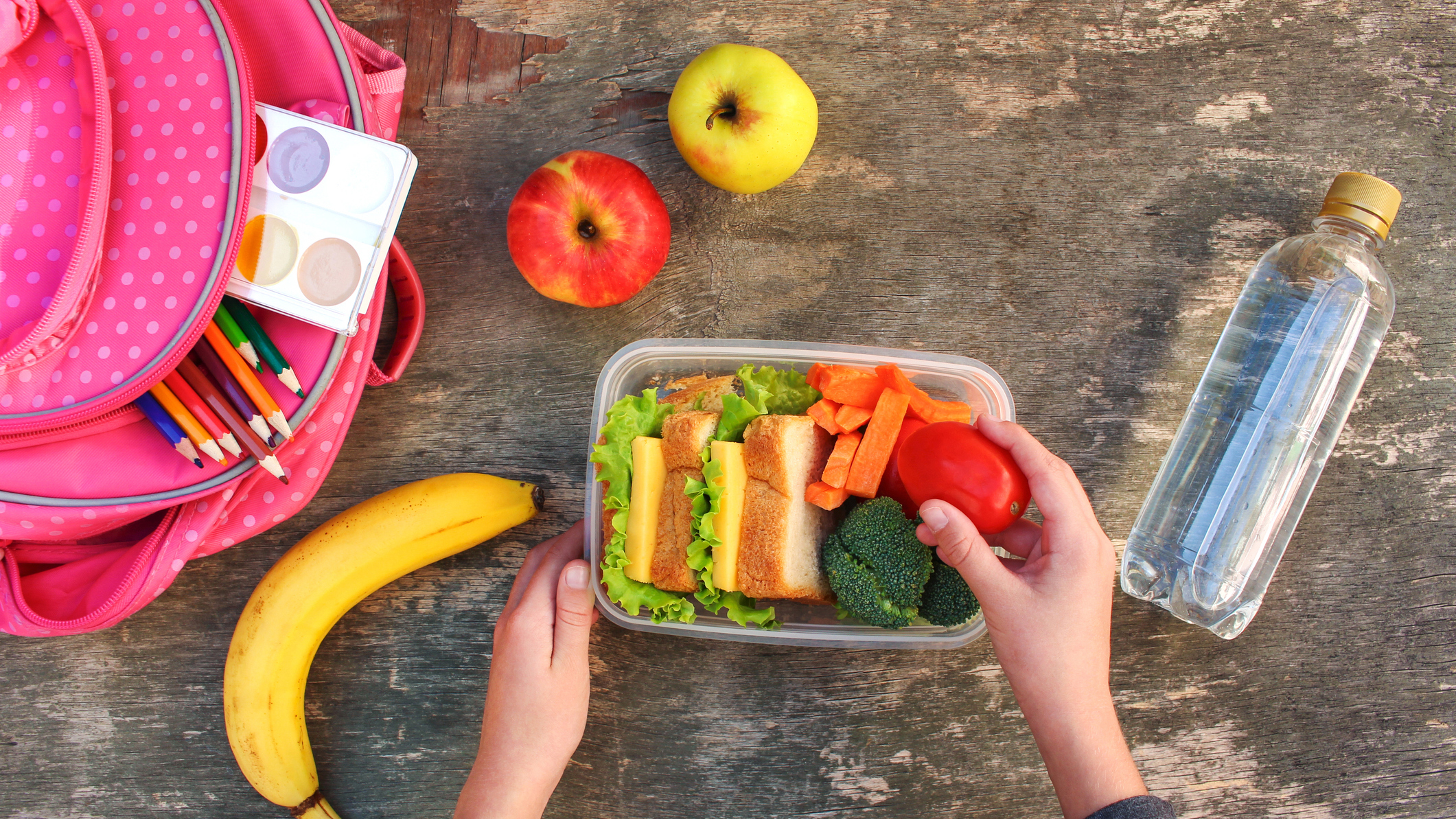 A képet felülről fotózták, látszik rajta egy iskolatáska, a hozzá való uzsonnástáska, és ami benne van. Szendvicsek, zöldségek és gyümölcsök.