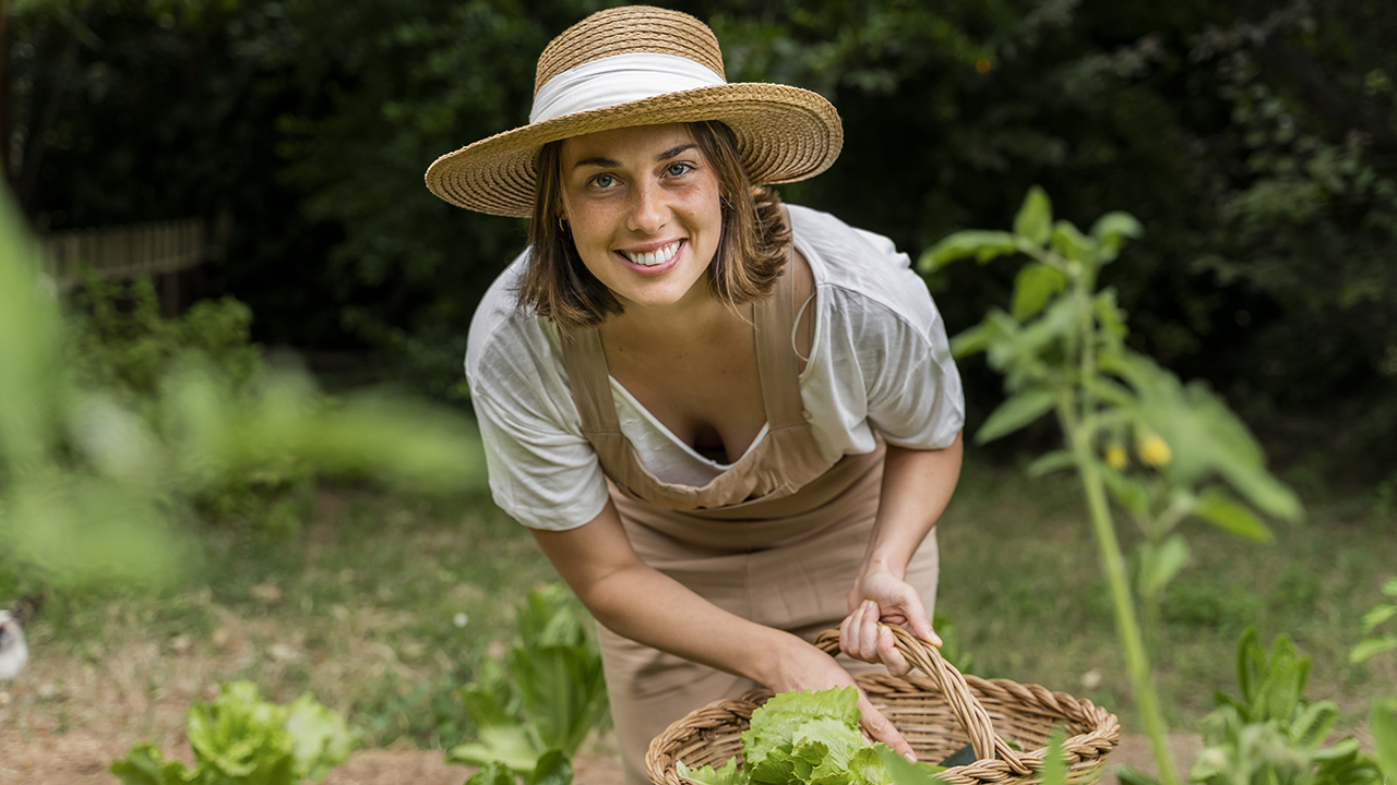 Egy fiatal nő a kamerába mosolyog, miközben egyik kezében kosarat tart a kertben guggolva.