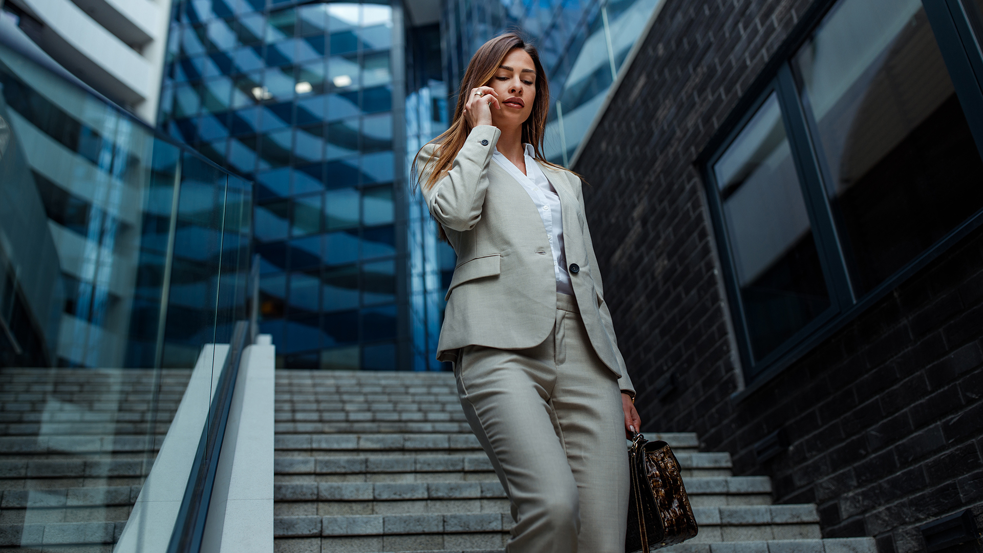 Egy fiatal nő sétál le a lépcsőn egy irodaház előtt zakóban és elegáns nadrágban, közben telefonál.