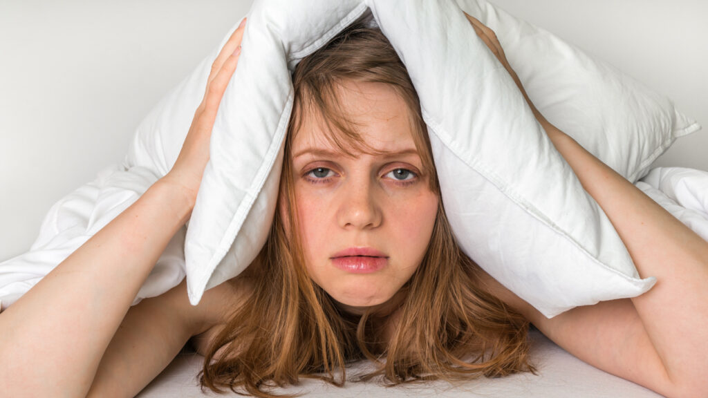 Young woman in bed covering ears with pillow because of noise