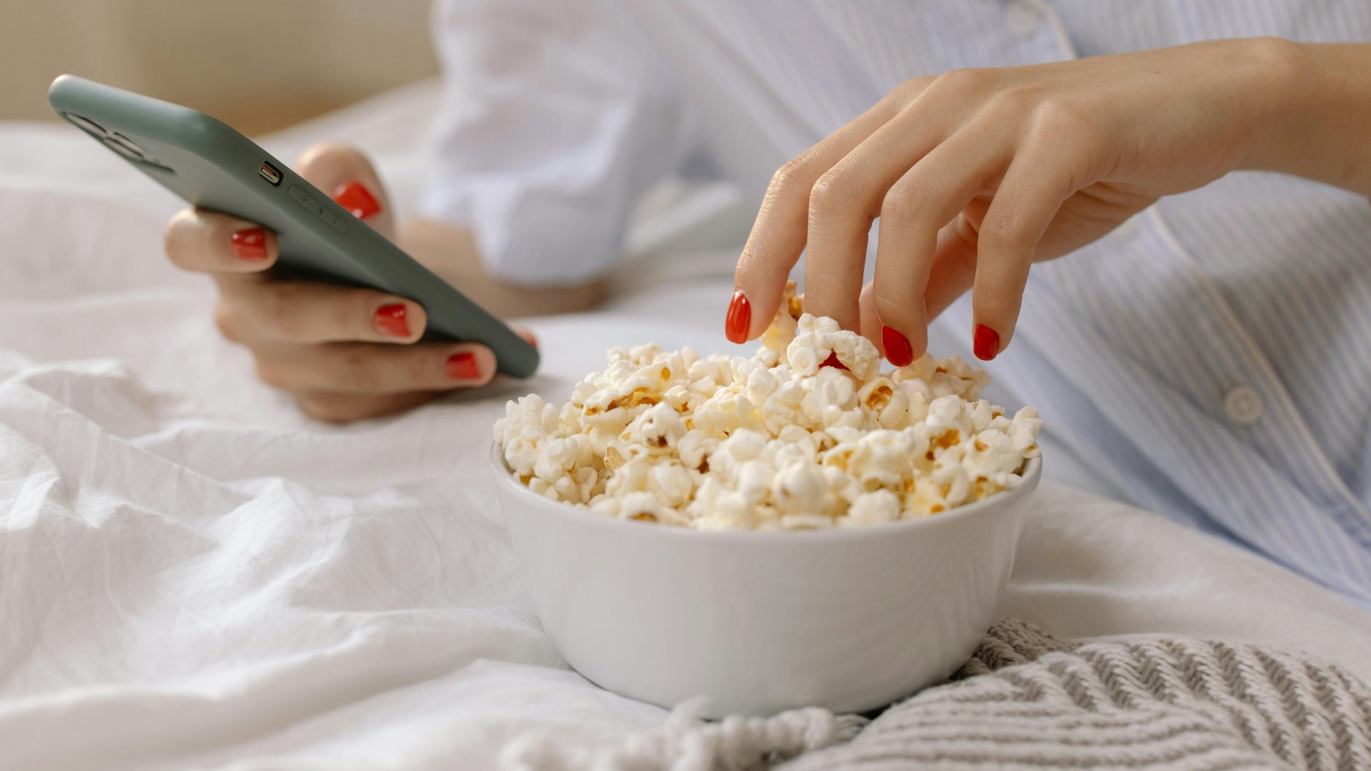 Egy nő az ágyon fekve telefonozik és popcornt eszik